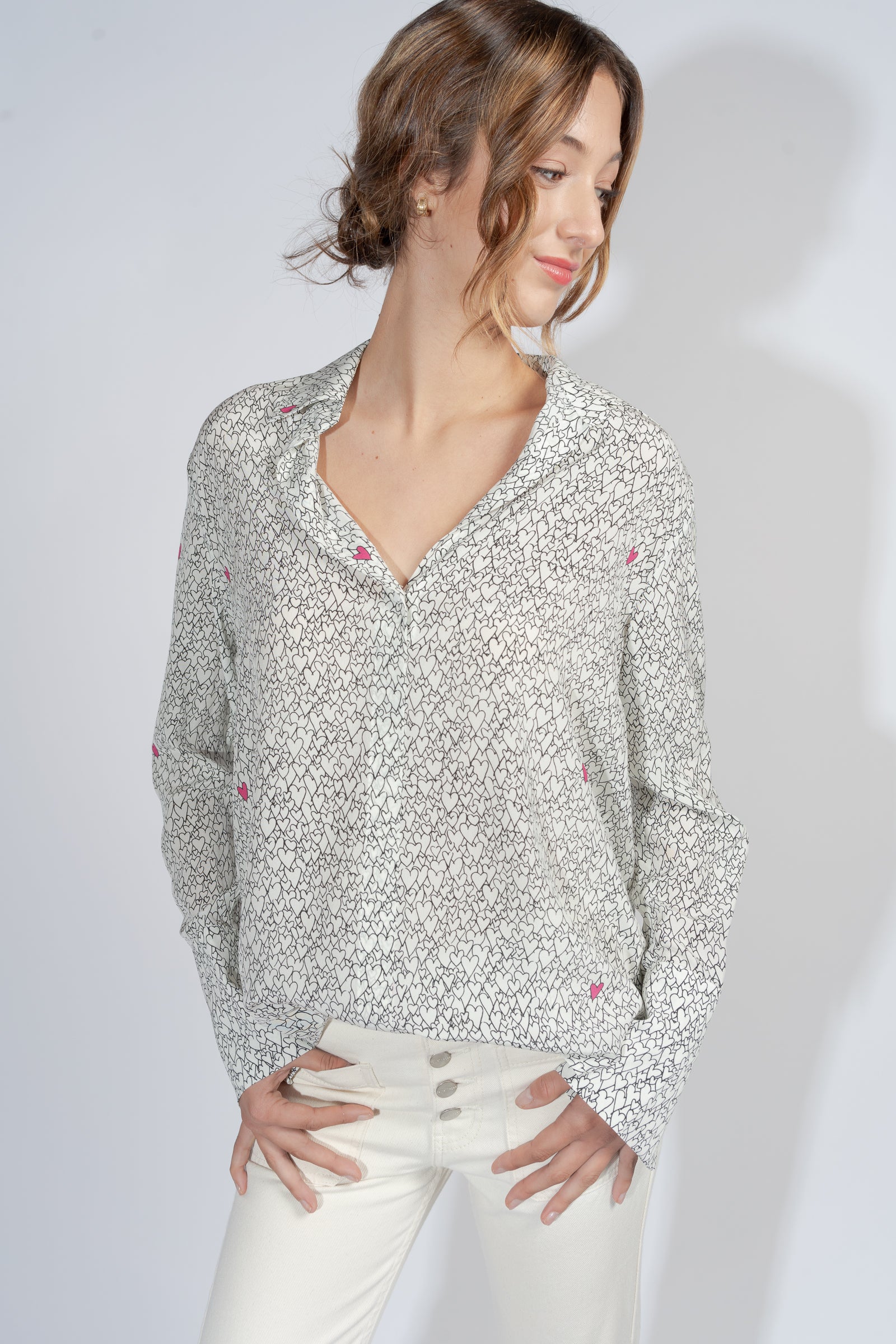 A woman wearing a Catherine Gee Daria French Cuff Silk-Heart Print blouse features white with a black pattern and small pink hearts, featuring a V-neck and long sleeves with French cuffs paired with high-waisted white pants with multiple buttons.