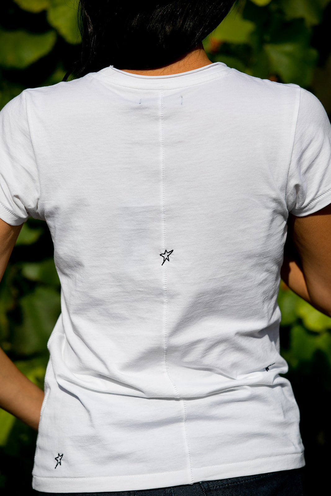 Back-view of a woman wearing a white Catherine Gee Embroidered Cotton Star t-shirt features a simple design with a small black embroidered star located in the center of the back, just below the shoulder blades, and another smaller star near the bottom left side. The t-shirt has a central seam running vertically down the back, adding a subtle design element on a blurred greenery background, highlighting the t-shirt embroidered.