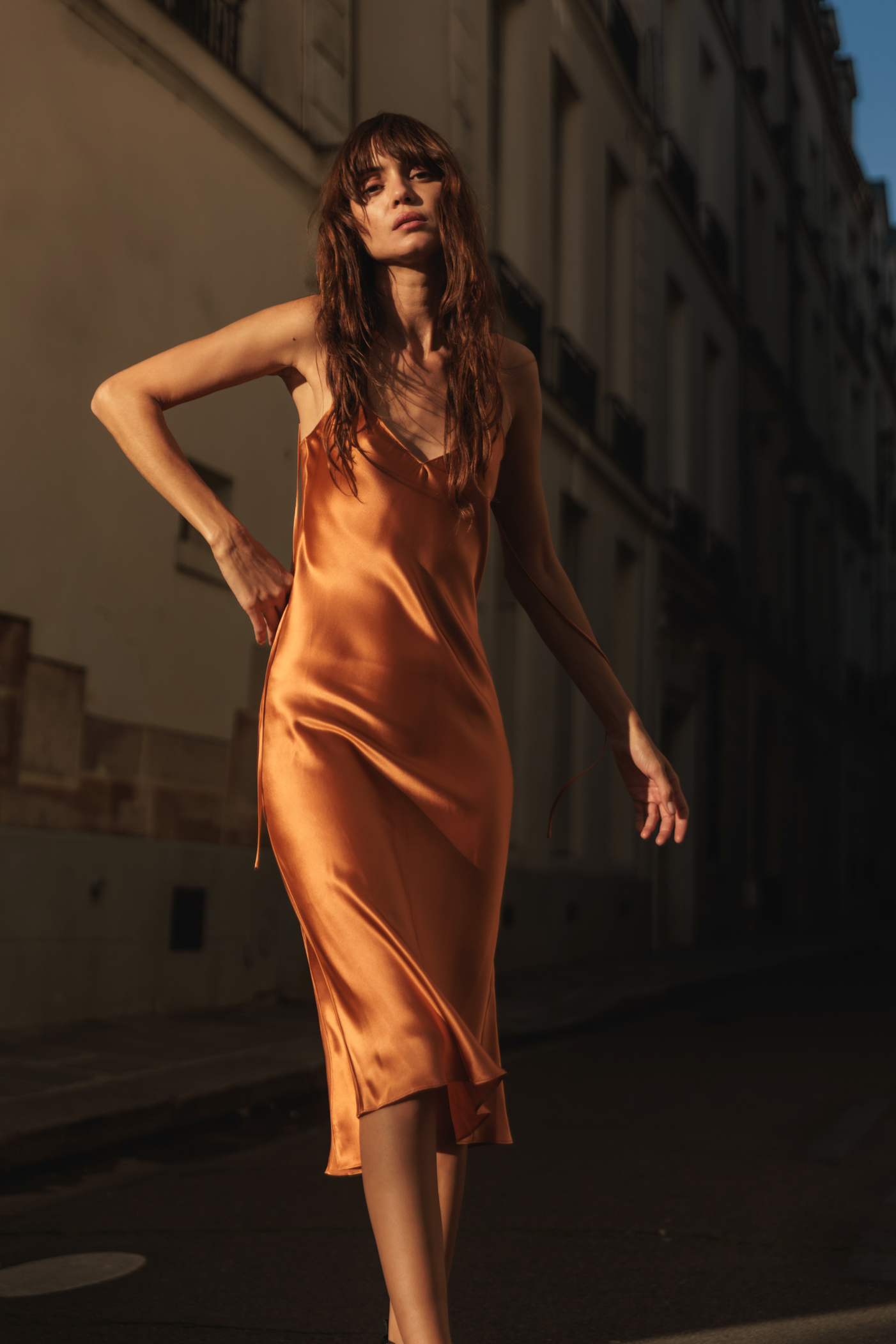 A woman wearing a Catherine Gee Emma Bronze Slip Dress. This elegant slip dress is made of silky, bronze-colored fabric with a slight sheen, and it falls below the knees features thin spaghetti straps and a V-neckline, creating a sophisticated and minimalist look. She is standing on a street with buildings in the background, illuminated by late afternoon lighting that casts long shadows, highlighting the luxurious fabric of the dress.