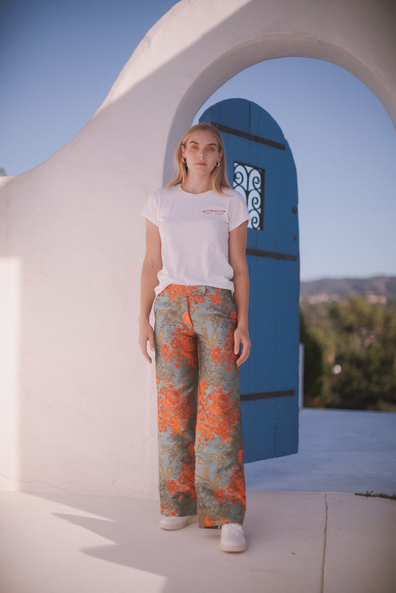 A woman standing in front of a white archway with a blue door in the background. She wears Catherine Gee Jacquard Stella Blue/Orange pants that feature a vibrant blue and orange floral pattern that stands out against the plain white T-shirt and the minimalist background. The setting appears to be outdoors, with a clear sky and some greenery visible in the distance, adding to the overall fresh and stylish look of the outfit.
