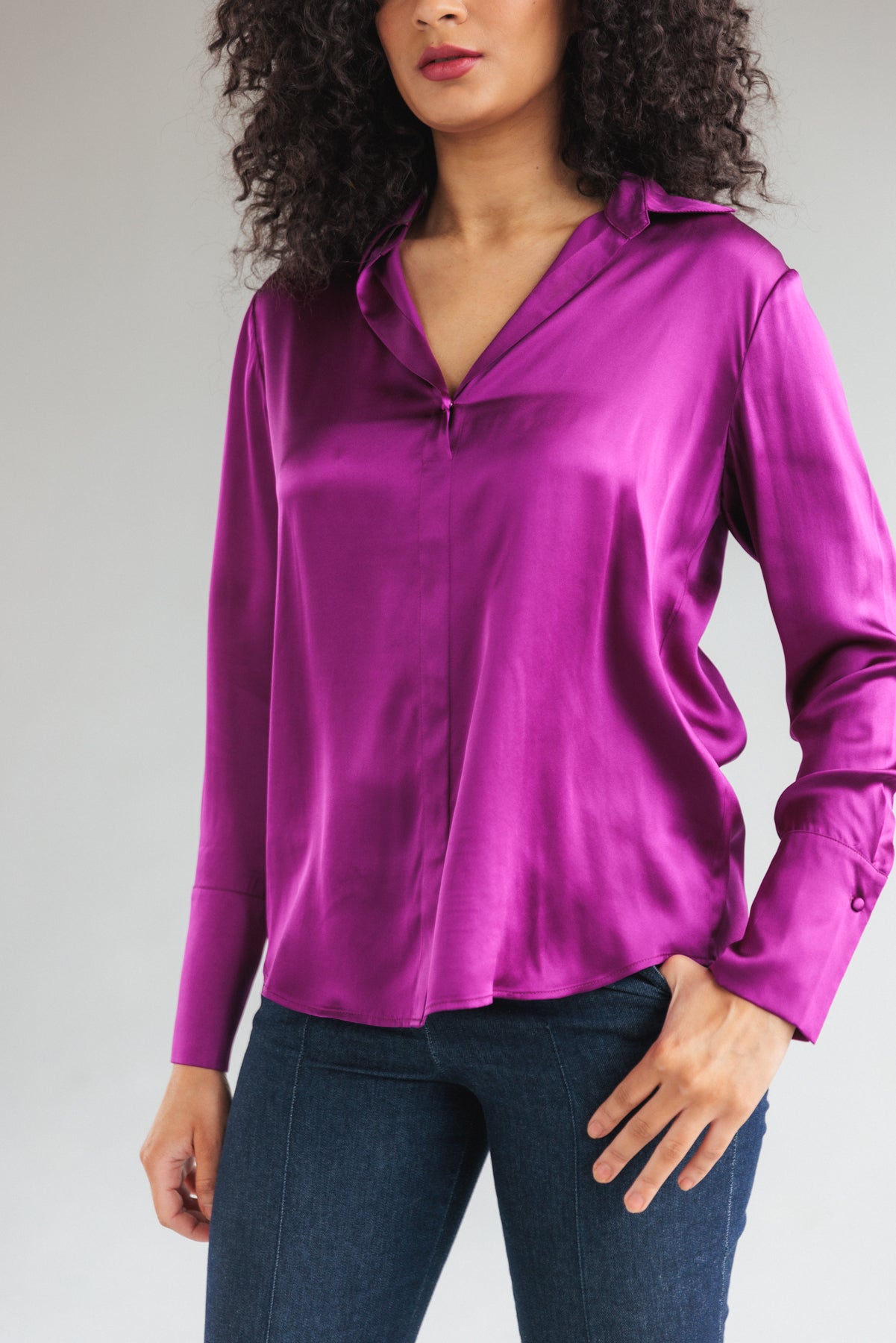 A woman wearing a Catherine Gee Daria French Cuff Silk Blouse Electric Orchid features a vibrant electric orchid-colored silk blouse with French cuffs, smooth, shiny texture, and a relaxed fit, paired with dark blue jeans.