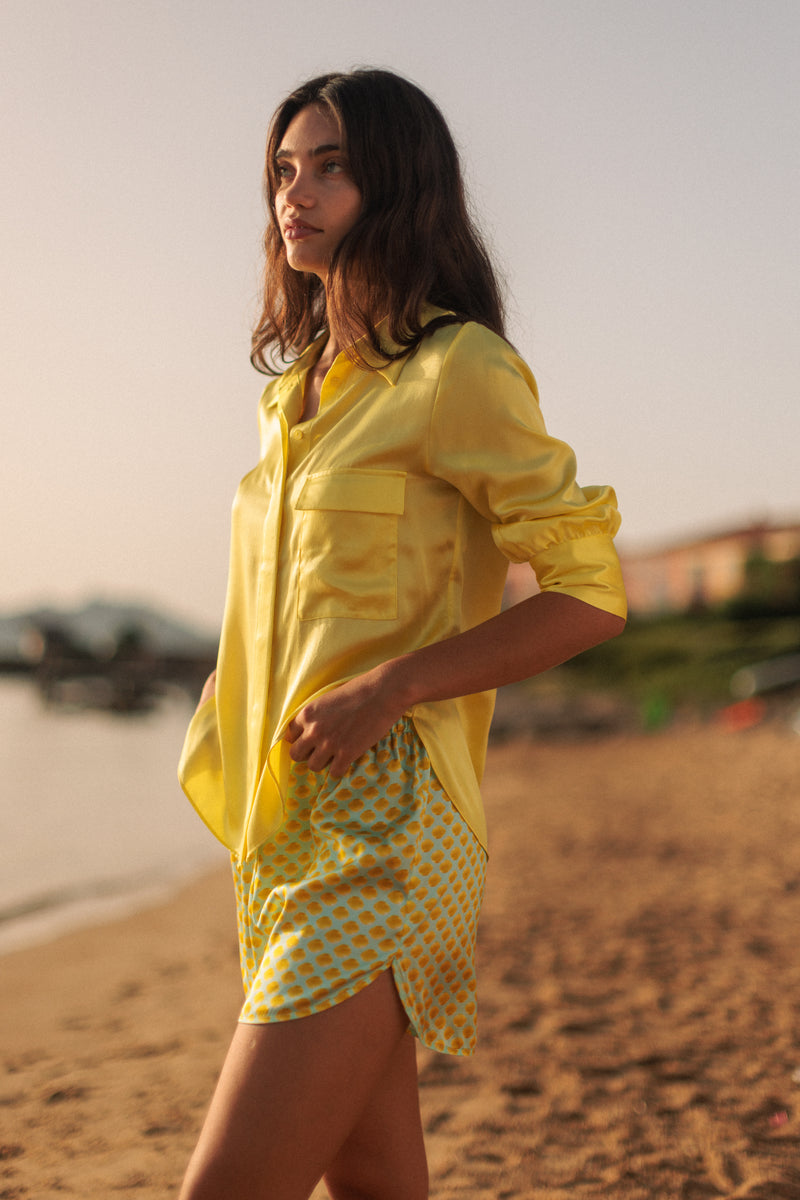 A woman standing on a beach, wearing a Catherine Gee Jamie Yellow Blouse. The blouse is a vibrant yellow color with a silky texture, featuring a button-down front and two chest pockets. The sleeves are rolled up to the elbows paired with yellow and white checkered shorts. The background includes a sandy beach, blurred buildings, and a calm body of water, suggesting a relaxed, summery setting.