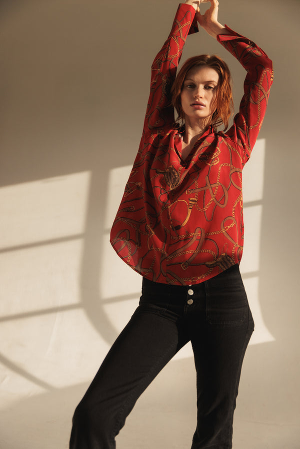 A woman wearing a Catherine Gee Daria French Cuff Silk Palermo blouse features a rich red color with an intricate pattern of ropes and chains in gold and brown hues giving the blouse a smooth and shiny texture. The blouse has long sleeves with French cuffs and a relaxed fit. She is also wearing high-waisted black pants with three visible buttons and has their arms raised above their head, with natural light streaming through the background which highlights the blouse's flowing design.