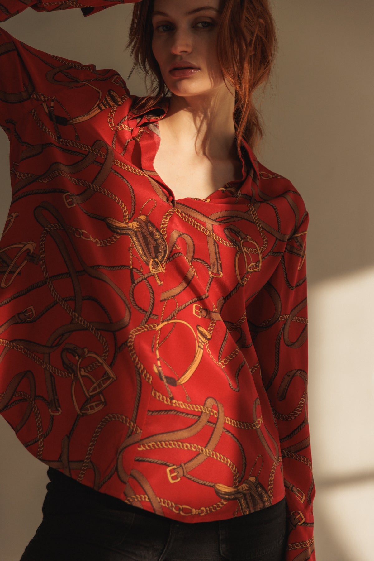 A woman wearing a Catherine Gee Daria French Cuff Silk Palermo blouse features predominantly red with an intricate pattern of ropes and buckles in shades of gold and brown, giving it a smooth and shiny texture. The blouse has a relaxed fit and features a French cuff design. She is posing with one arm raised, which highlights the flow and drape of the blouse emphasizing the luxurious quality of the silk material.