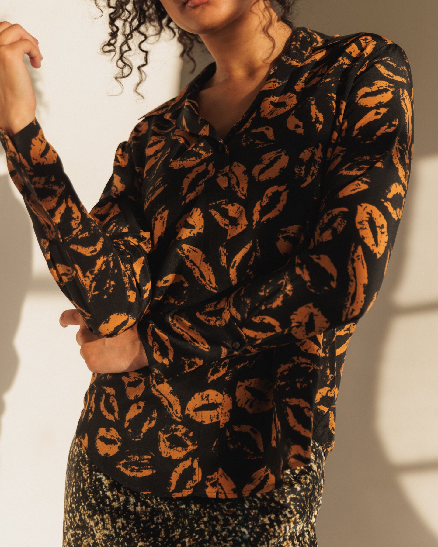 A woman wearing a Catherine Gee Daria French Cuff Silk-Mocha Lips blouse featuring a black background with an all-over print of mocha-colored lips, giving it a bold and unique look. The fabric appears to be silk, providing a smooth and slightly shiny texture features a collar, long sleeves with French cuffs, and a relaxed fit. She is standing with one arm bent and the other arm resting on it, creating a casual and relaxed pose.