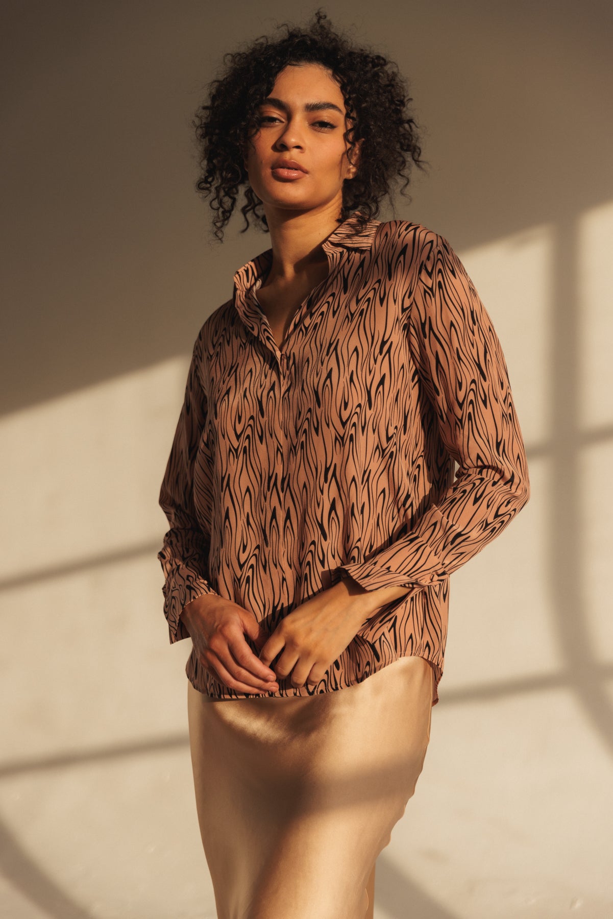 A woman wearing a Catherine Gee Daria French Cuff Silk Wishbone blouse features a distinctive wishbone pattern in a warm, earthy tone. The fabric appears to be lightweight and silky, draping elegantly over the person's body, and has a classic collar and long sleeves with French cuffs, adding a touch of sophistication paired with a light-colored, satin-like skirt that complements the blouse. The overall look is stylish and refined, suitable for both casual and formal occasions.