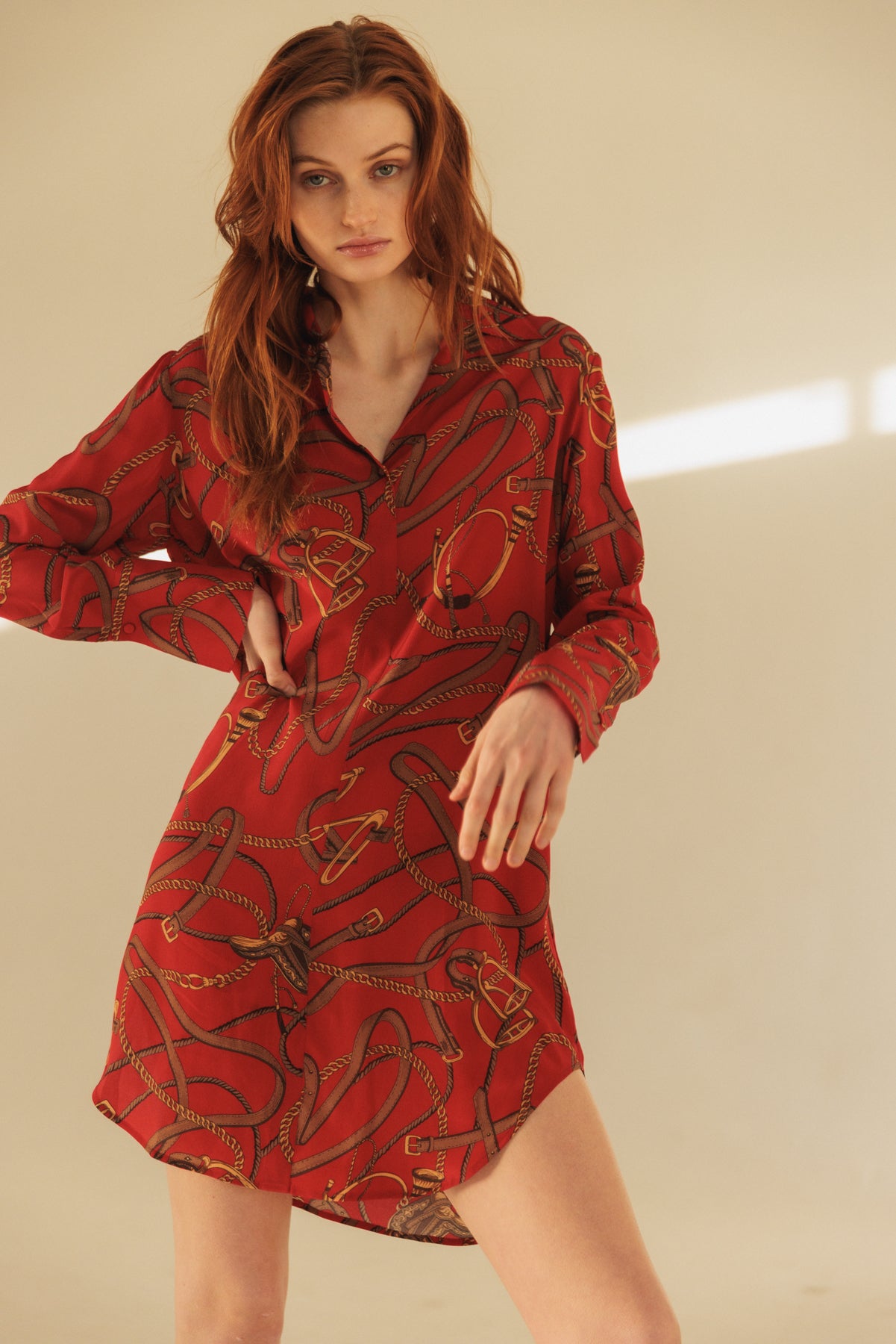 A woman wearing a Catherine Gee Daria Silk Shirt Palermo Dress. This vibrant red dress features an intricate pattern of intertwined ropes and chains in gold and black, has long sleeves, and a slightly loose fit, with a hem that is shorter in the front and longer in the back. The neutral beige background highlights the dress's bold colors and detailed design, making it a standout piece suitable for various occasions.