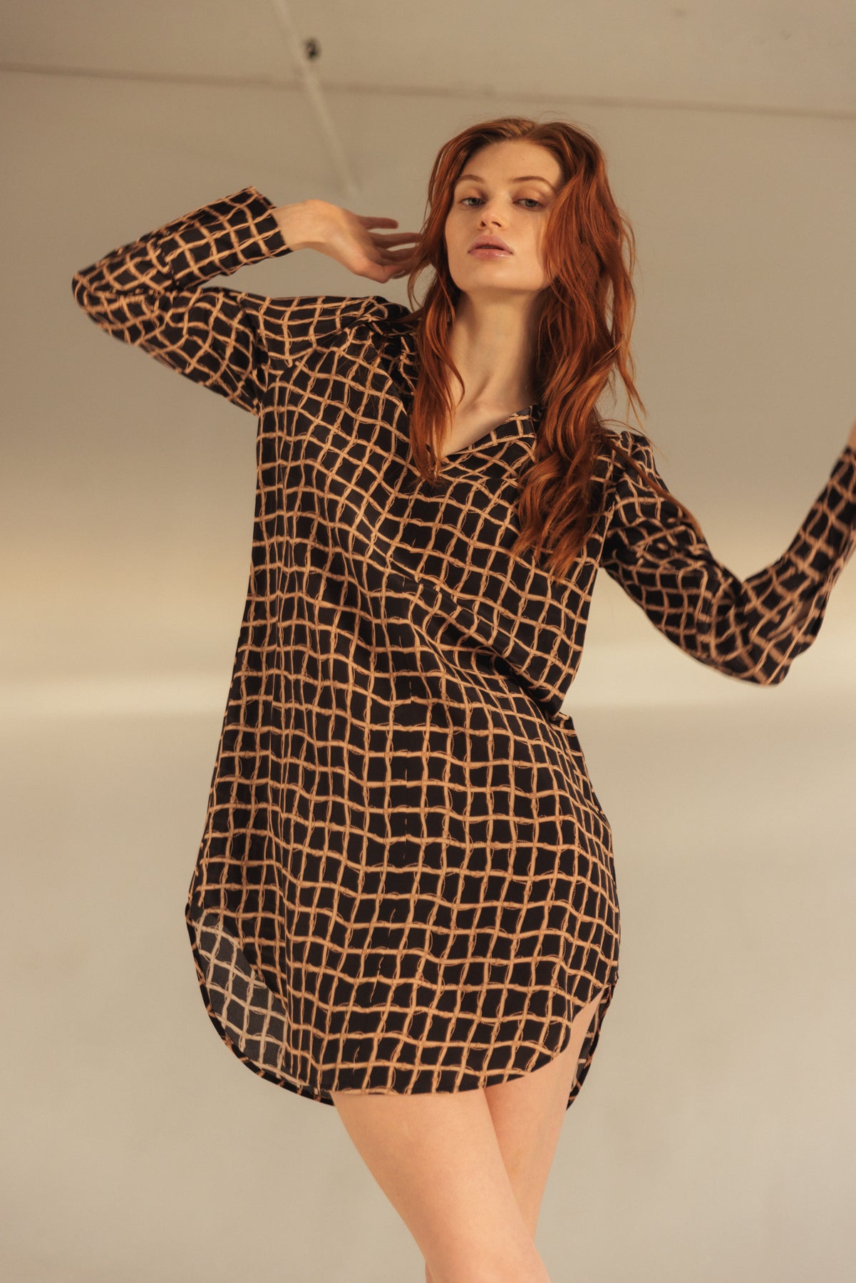 A woman wearing a Catherine Gee Daria Recoleta dress features a black base with a grid-like pattern of beige lines. It has a loose, relaxed fit with long sleeves and a slightly curved hemline that falls above the knees. The lightweight, flowy fabric adds an effortless elegance, making it suitable for casual or semi-formal occasions, and perfectly combines comfort and style.