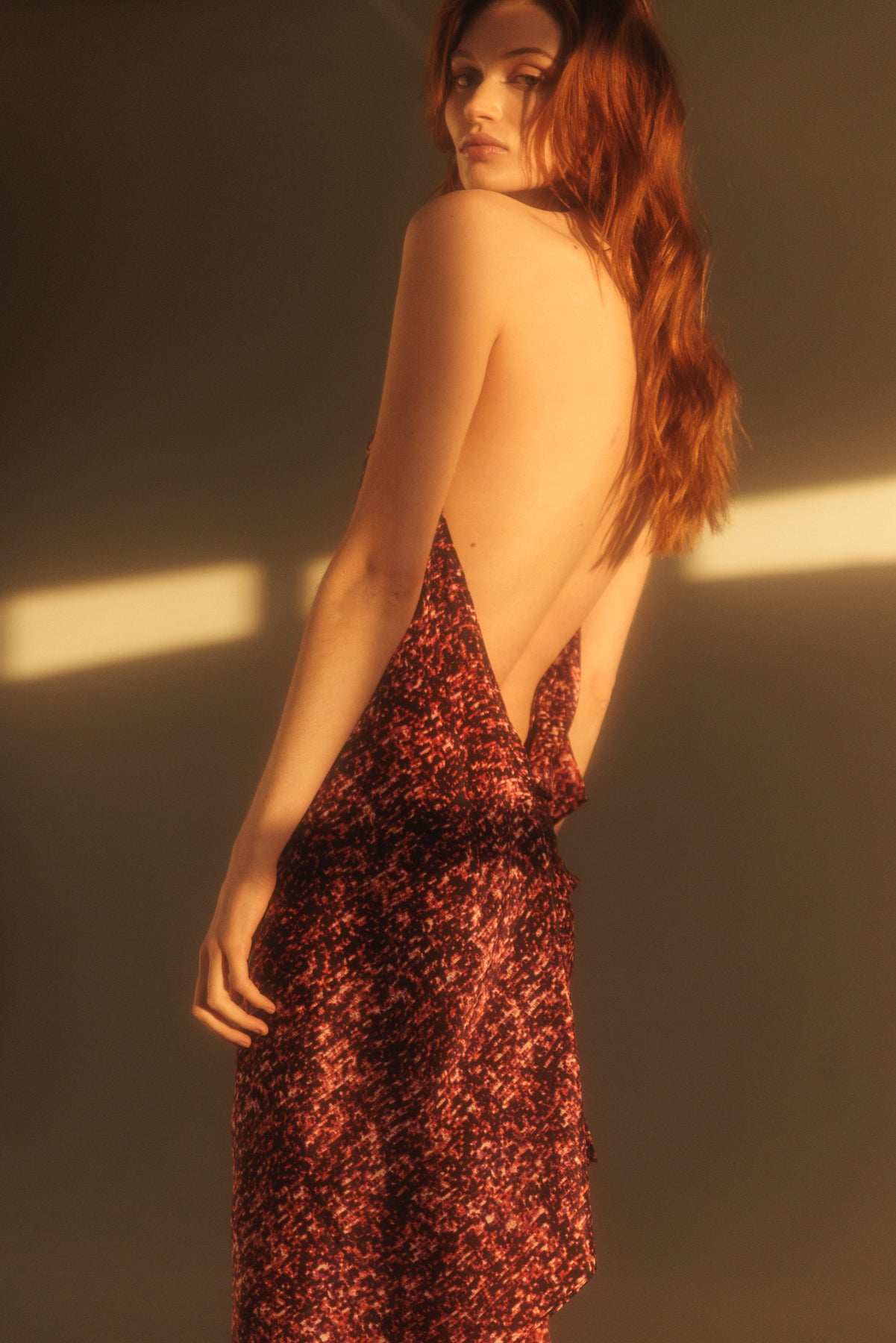 A woman wearing a Catherine Gee Diane Red Sequin dress features a deep, plunging back, revealing much of the wearer's back. The fabric is red with a sequin pattern, creating a shimmering effect. The lighting in the image casts a warm glow, highlighting the texture and color of the dress. The overall aesthetic is elegant and sophisticated, making the dress stand out as a statement piece.