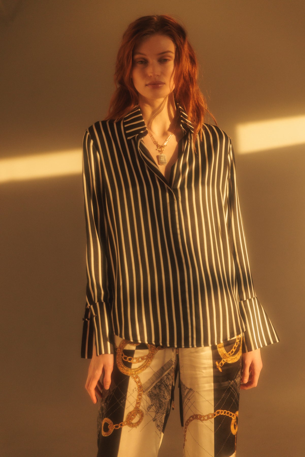 A woman wearing a Catherine Gee Daria French Cuff Silk black and white stripes blouse features a classic collar and long sleeves with French cuffs. She is also wearing patterned pants with chain and geometric designs. The lighting in the image creates a warm, golden hue, adding a stylish and sophisticated ambiance to the overall look.