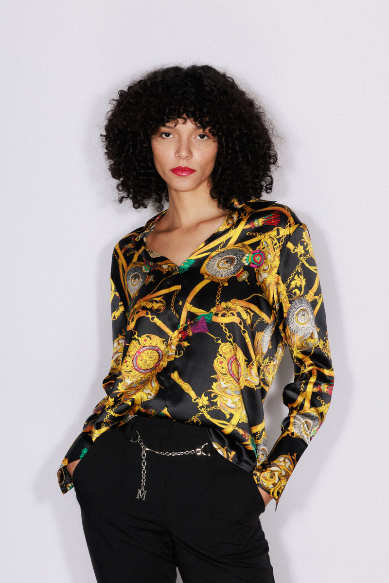 A woman wearing a Catherine Gee Daria French Cuff Silk Versailles blouse features a luxurious black base with intricate gold and colorful chain and medallion patterns. It has a deep V-neckline and long sleeves with French cuffs, paired with black pants and a chain belt with a pendant. The overall look is elegant and stylish, highlighting the rich design of the blouse.