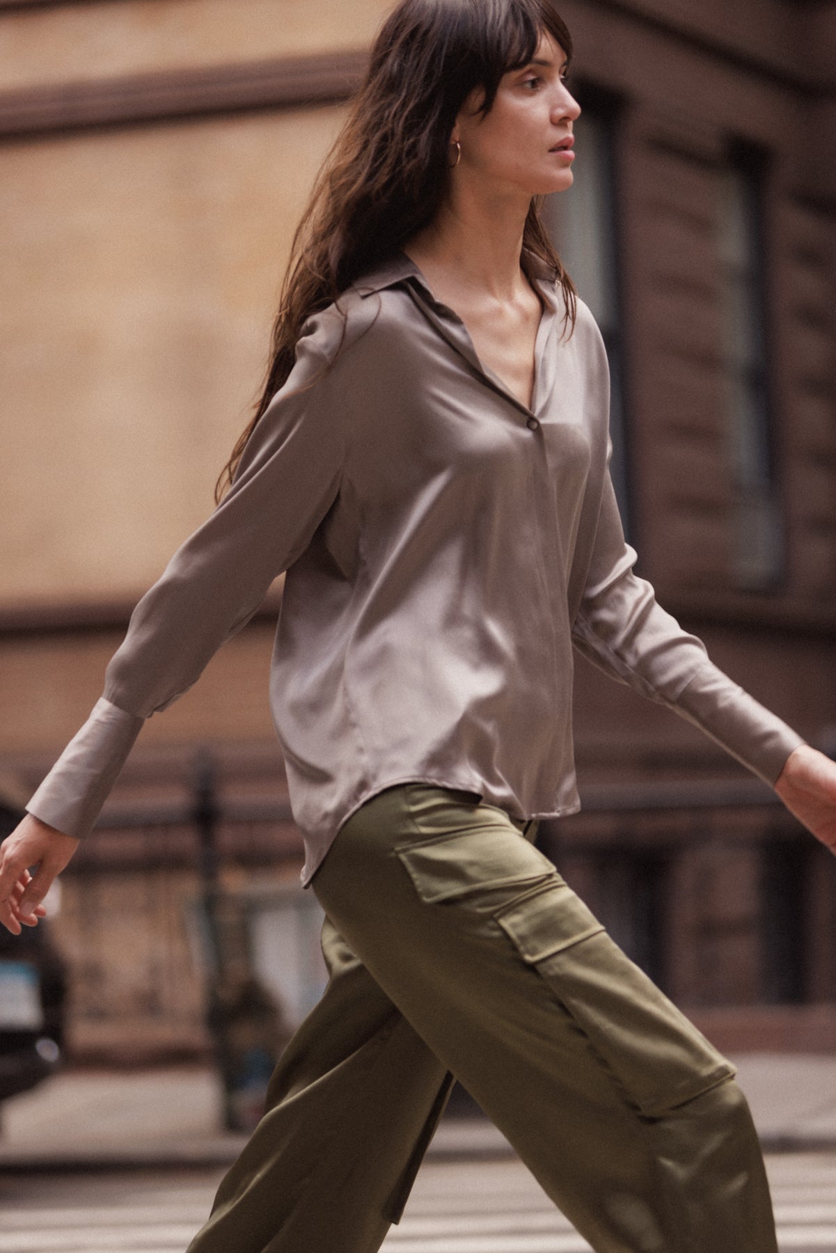 Woman wearing a Catherine Gee Daria French Cuff Silk-Grey blouse paired with olive green cargo pants. The blouse is loose-fitting with a V-neck and long sleeves, while the pants have large pockets on the sides. The background shows an urban setting with blurred buildings and a sidewalk.