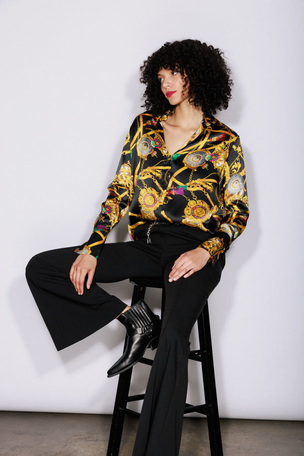 A woman sitting on a stool, wearing a Catherine Gee Daria French Cuff Silk Versailles blouse features a luxurious and intricate design with a black base and ornate gold and colorful chain and medallion patterns. She also wears black flared pants and black ankle boots, creating a sophisticated and stylish look on a plain white background that highlights the outfit.