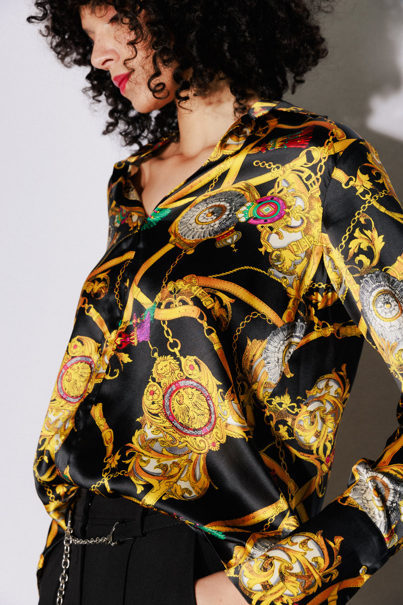 A Catherine Gee Daria French Cuff Silk Versailles blouse features a luxurious black silk fabric decorated with an intricate and vibrant print of gold chains, medallions, and ornate designs. The pattern includes elements of red, green, and blue, adding to its opulent appearance. The blouse has a relaxed fit with a deep V-neckline and long sleeves, making it a stylish and elegant piece suitable for various occasions.