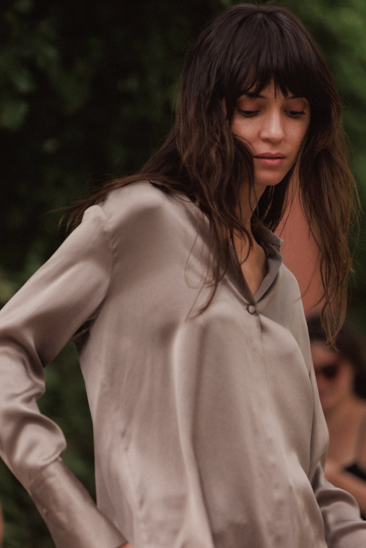 A woman wearing a Catherine Gee Daria French Cuff Silk-Grey blouse is made of smooth, shiny silk and features a V-neckline with a single button closure. The sleeves are long with French cuffs, and the blouse has a relaxed, elegant fit. The background is blurred with greenery.