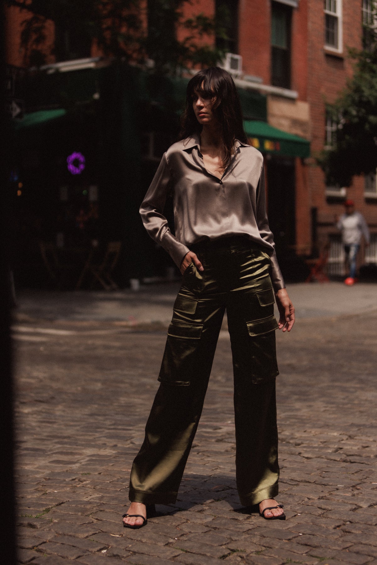 A woman is standing on a cobblestone street wearing Army Green color Catherine Gee Cargo Pants. The pants are high-waisted with multiple pockets and have a loose, wide-leg fit paired with a silky, long-sleeved blouse in a neutral color and black sandals. The background shows an urban setting with buildings and some greenery.