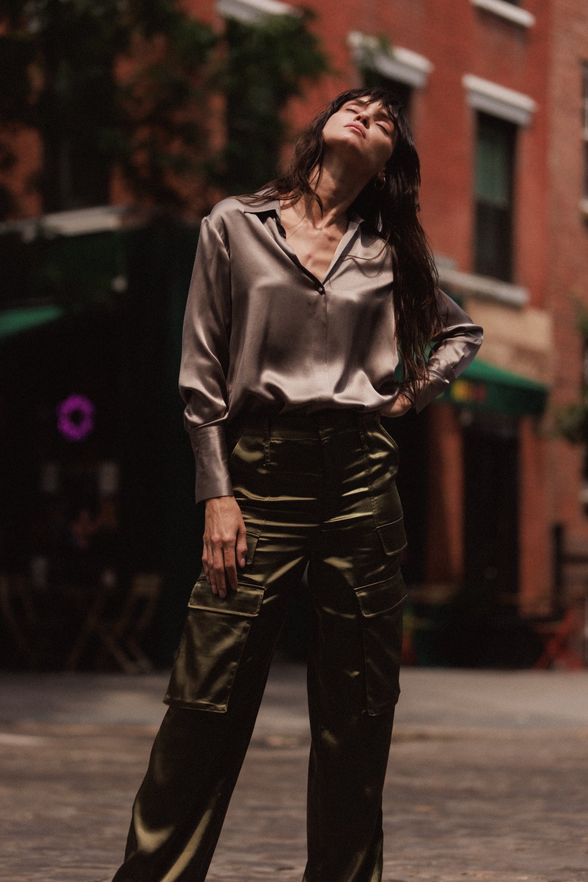 A woman wearing a Catherine Gee Daria French Cuff Silk-Grey blouse paired with high-waisted, olive green cargo pants. The blouse has a shiny, smooth texture and a loose fit, while the pants have multiple pockets and a structured design. The background shows an urban setting with brick buildings and some greenery highlighting the stylish and modern, combining elegance with a touch of utility.