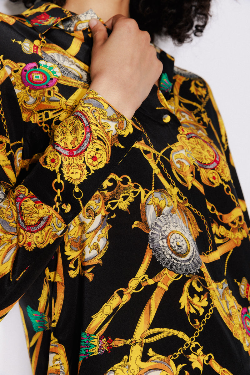 Close-up of a woman holding the collar of the dress with one hand wearing a Catherine Gee Versailles - Daria Dress features an intricate design with gold chains, medallions, and ornate patterns on a black background, showcasing the detailed and luxurious fabric design.