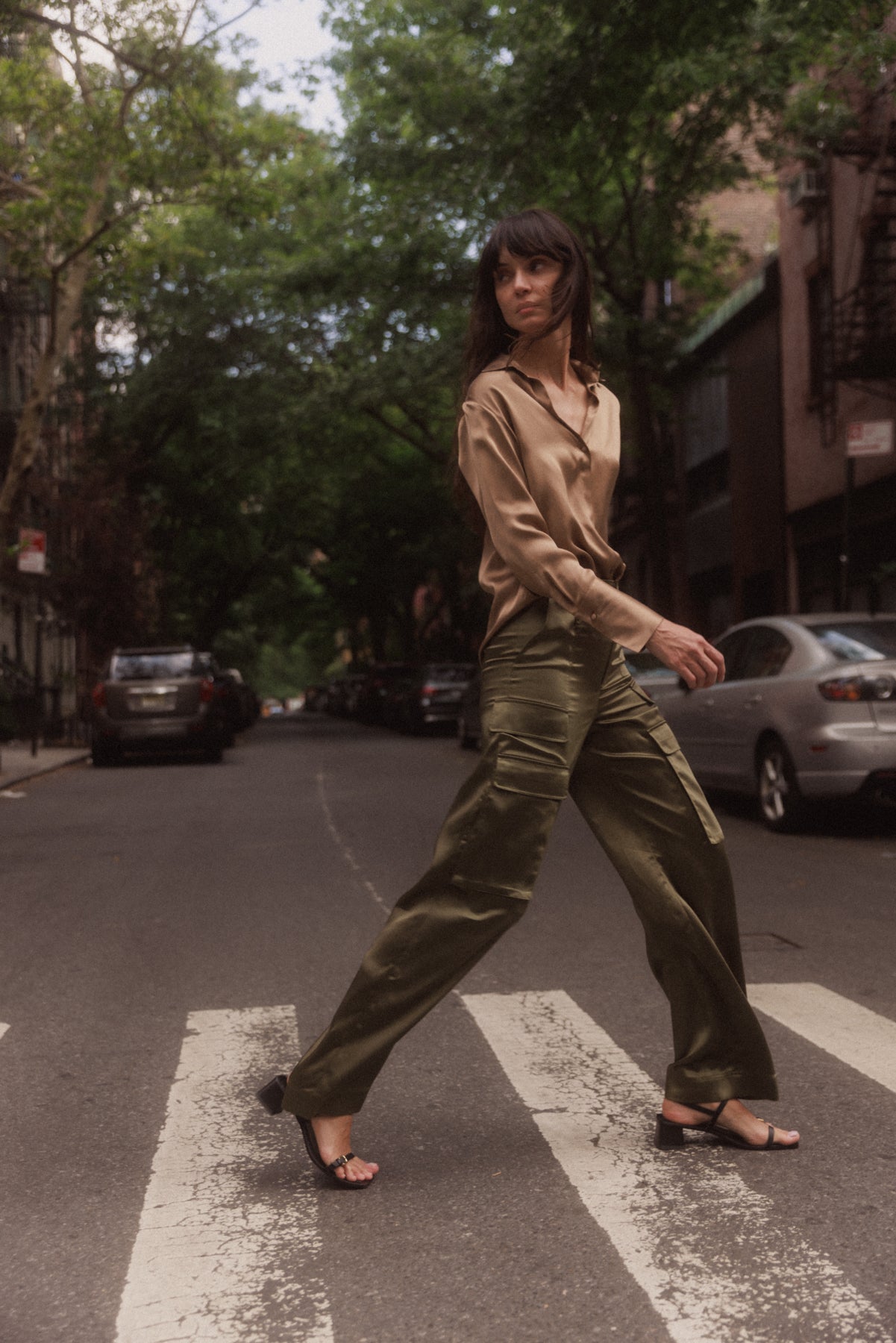 A woman walking across a street in an urban setting, wearing Army Green color Catherine Gee Cargo Pants paired with a light brown blouse and black sandals. The background features parked cars and trees lining the street, creating a serene and stylish city scene. 