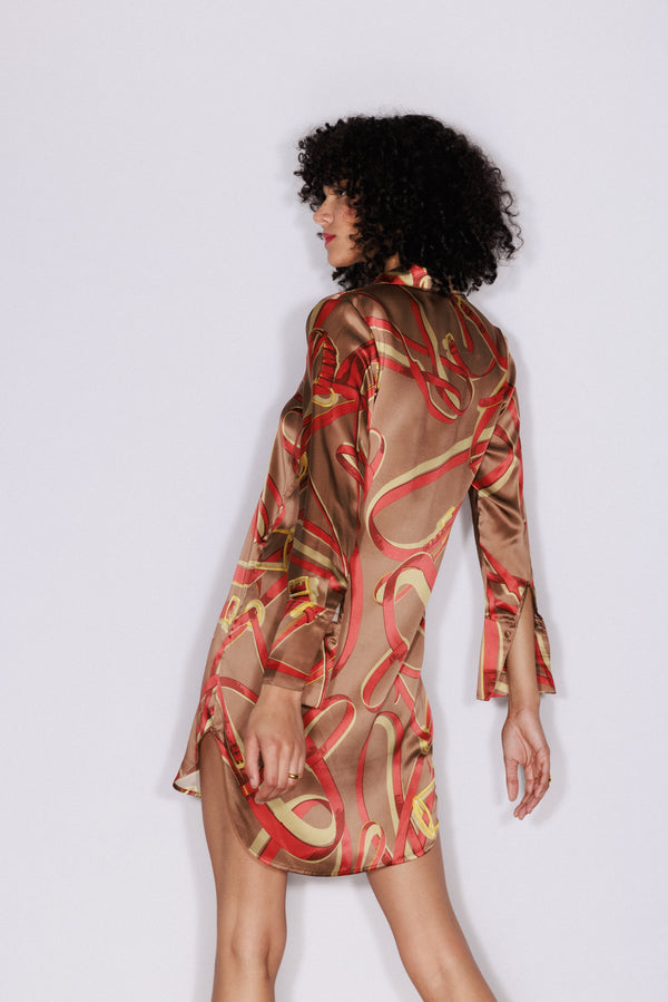 A woman wearing a Catherine Gee Daria Dress Lexington Equestrian pattern features a silky fabric with a brown base color and an abstract design of red and yellow swirls. The dress has long sleeves with slits near the cuffs and a short hemline that falls above the knees. She is posed with one arm slightly bent and the other hanging down, with her back facing the camera.