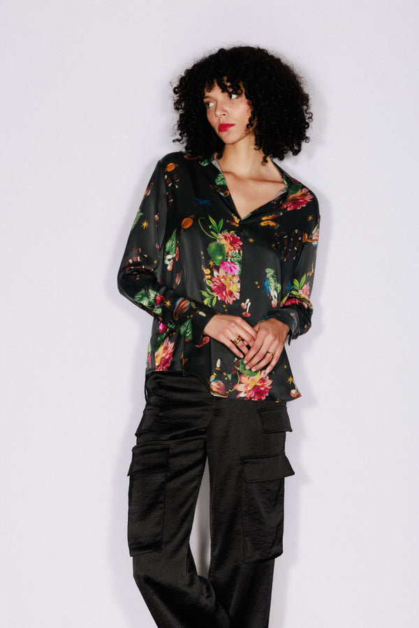 A woman wearing a Catherine Gee Daria French Cuff Silk Serpentine blouse features a black base with a vibrant eclectic print featuring various elements such as flowers, mushrooms, snakes, butterflies, and planets. It has a relaxed fit, V-neckline, and French cuffs, adding a touch of elegance paired with black cargo pants with multiple pockets. The outfit blends luxury and casual styles, creating a unique and fashionable look.