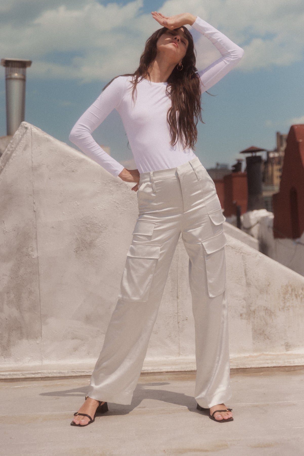 A woman wearing a white long-sleeve top and silver cargo pants by Catherine Gee stands on a rooftop with a cityscape in the background. She is posing with one hand on the hip and the other shielding their eyes from the sun. The pants have multiple pockets and a shiny texture, paired with black open-toe sandals. The sky is clear with a few clouds, adding to the bright and sunny atmosphere.