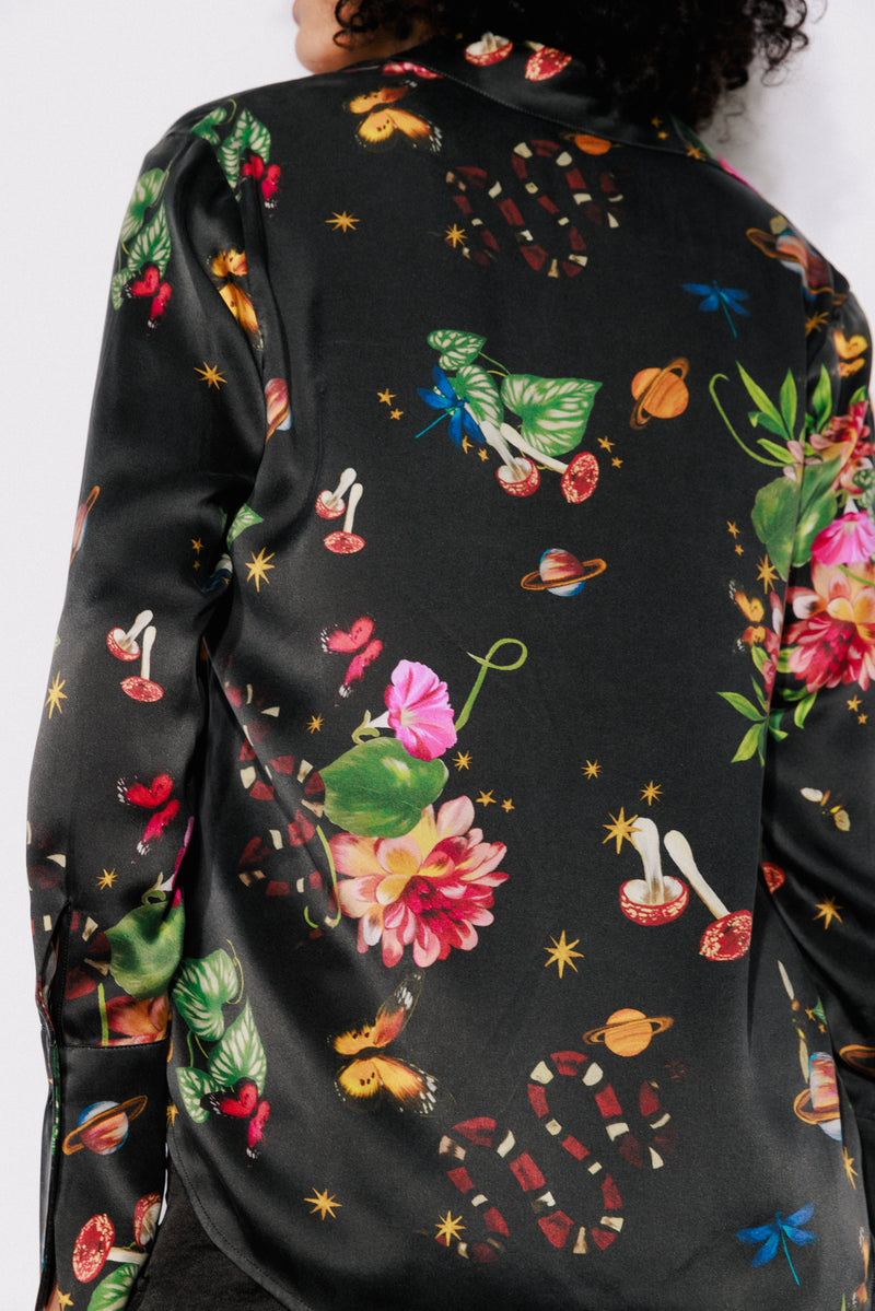 Back-view of a Catherine Gee Daria French Cuff Silk Serpentine blouse features black with a vibrant, eclectic print featuring various elements such as flowers, mushrooms, snakes, butterflies, and planets. The design is intricate and colorful, making the blouse visually striking and unique. The silk material gives it a luxurious and smooth appearance.