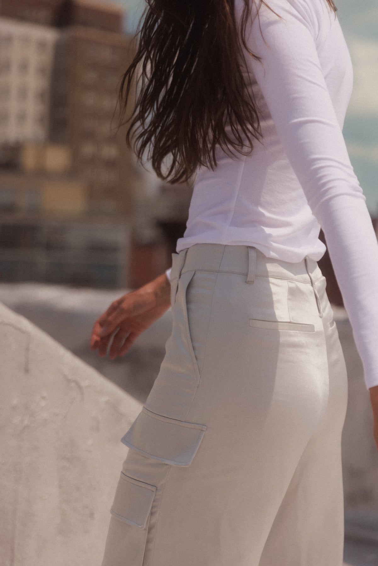 A woman wearing Catherine Gee Cargo Silver Pants and a white long-sleeve shirt standing outdoors. The pants have multiple pockets and a fitted waist. The background shows an urban setting with blurred buildings.