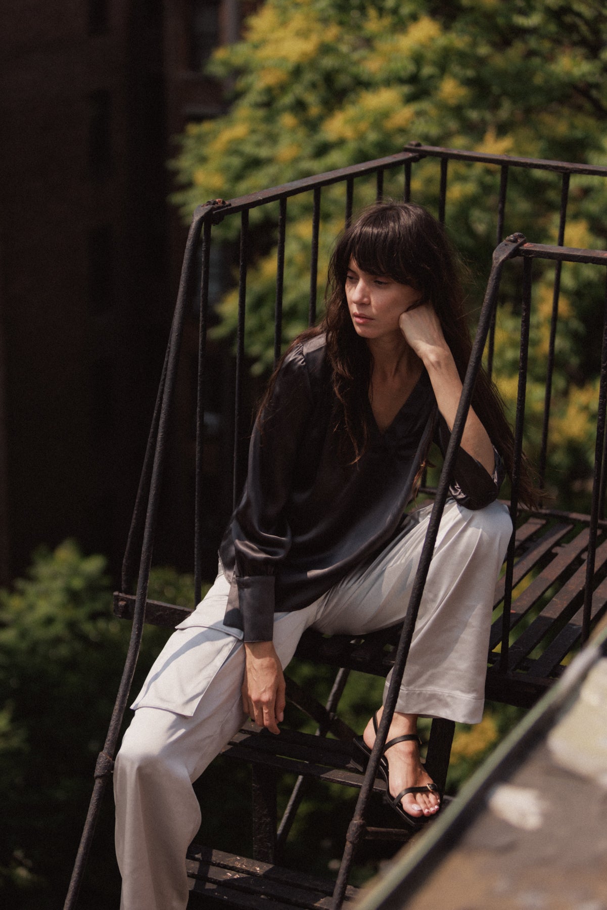 A woman is sitting on an outdoor metal staircase, wearing a dark long-sleeved top and silver-colored cargo pants. The background features greenery and a blurred building highlights the Catherine Gee Cargo Silver Pants in a casual, urban setting.