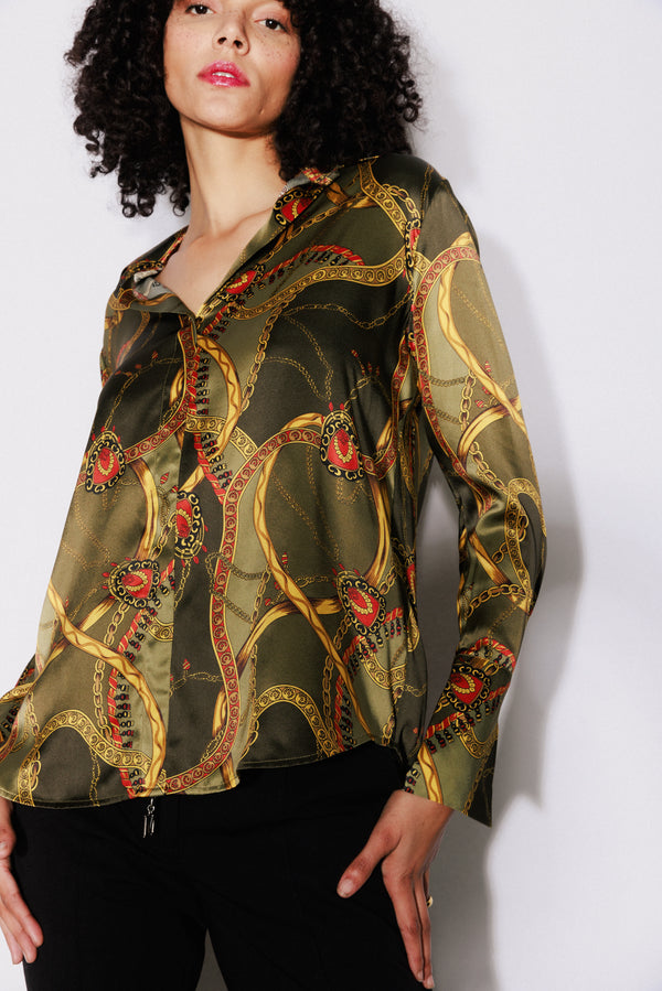 A woman wearing a Catherine Gee Daria French Cuff Silk Olive Equestrian blouse features an olive green color with an intricate equestrian-themed print, including detailed chain patterns and red accents paired with black pants creating a stylish chic look. Made of luxurious silk, the blouse has a button-down front and French cuffs, adding to its sophisticated appearance. 