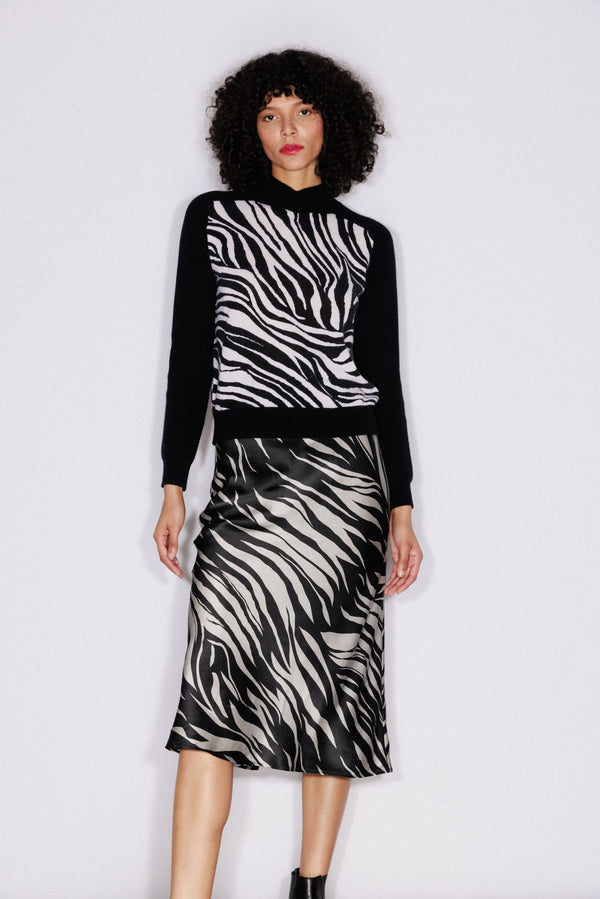 A woman wearing the Catherine Gee Gwen Color Block Cashmere Zebra Sweater. This luxurious sweater features a striking zebra pattern on the front, with bold black and white stripes creating a dramatic look. The sleeves and back are solid black, providing a chic contrast. Made from high-quality cashmere, the sweater offers both comfort and style paired with a zebra-print skirt, this outfit is perfect for fashion enthusiasts seeking unique and trendy pieces.