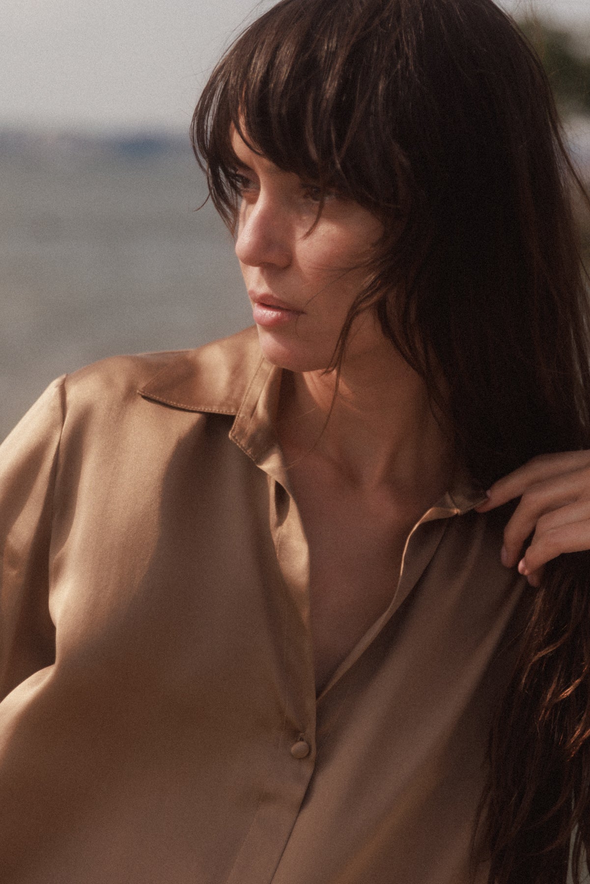 A woman with long hair wearing a Catherine Gee Daria French Cuff Silk-Mocha blouse, made of luxurious silk fabric with a smooth, shiny finish, featuring a classic collar, long sleeves with French cuffs, and a relaxed fit. The background suggests a sunset in an outdoor setting, possibly near a body of water giving a warm, calm atmosphere, adding a peaceful vibe.