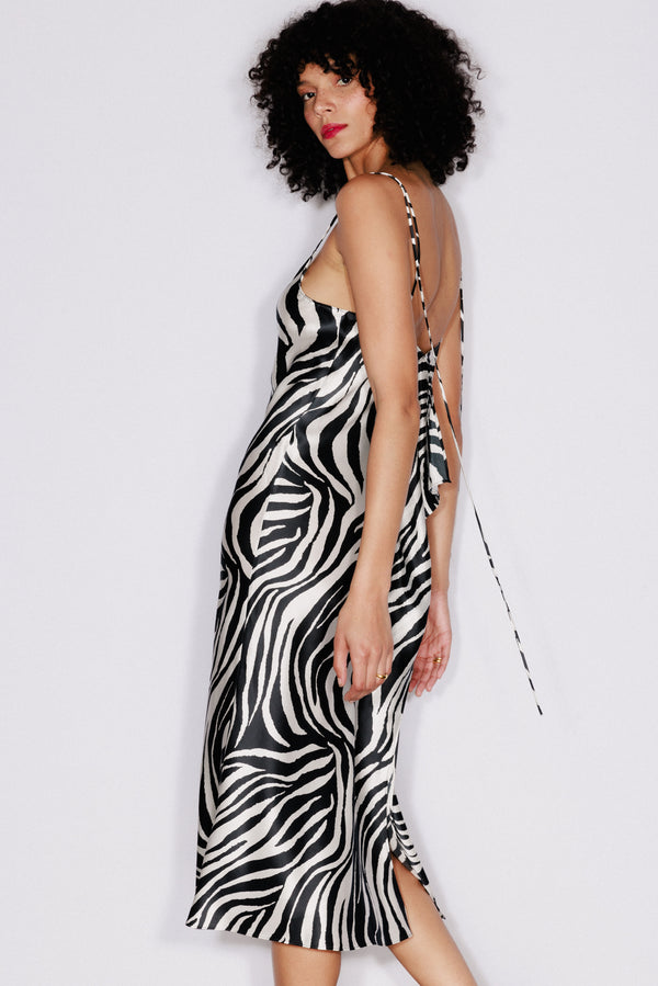 A woman wearing a Catherine Gee Emma Zebra Slip dress features a black and white zebra print pattern and has a midi length. It has thin spaghetti straps that crisscross at the back, creating an open-back design. The fabric appears to be silky and smooth, draping elegantly over the body. The dress also has a small slit at the hem, adding a subtle touch of charm.