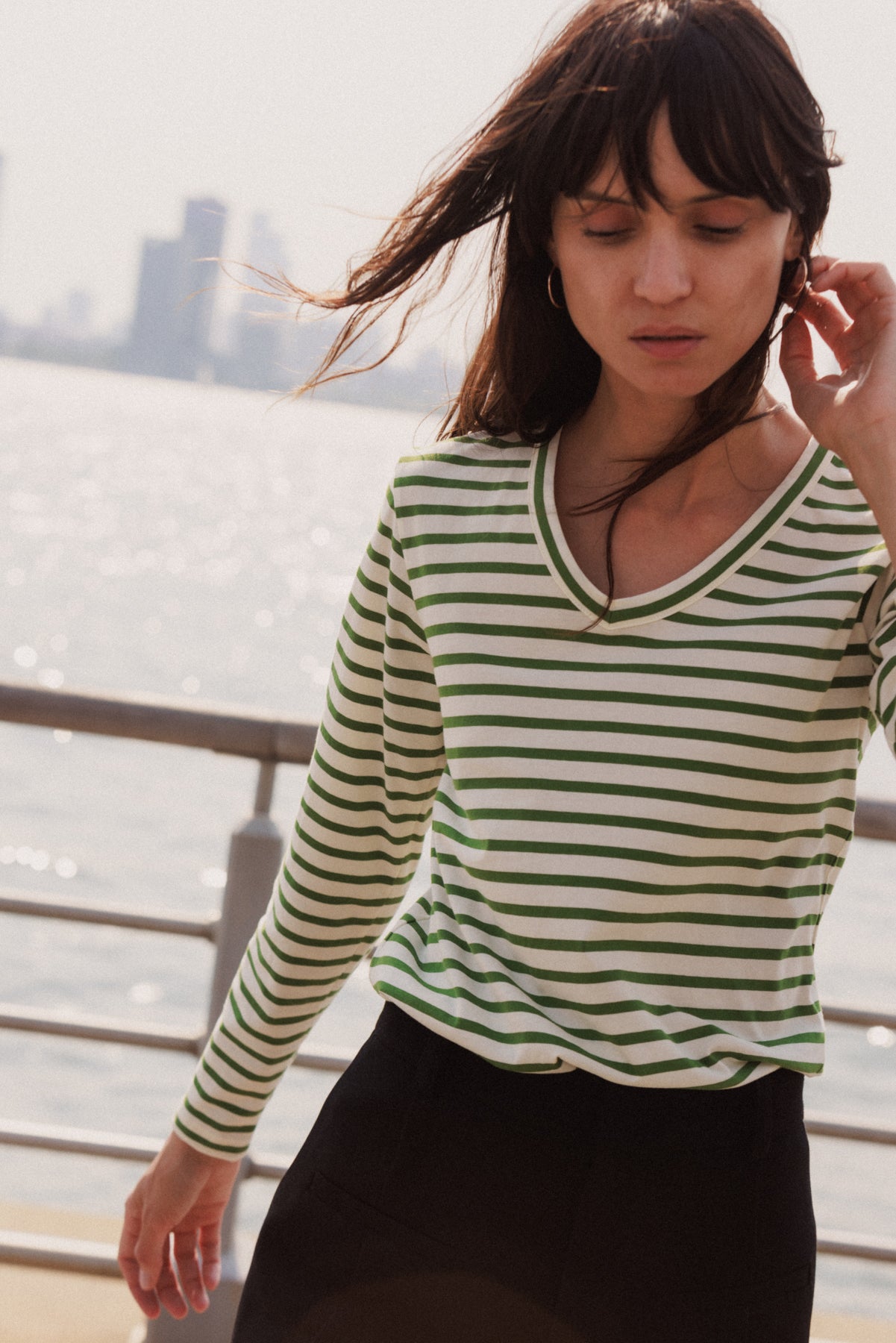 A woman standing outdoors near a waterfront with a city skyline in the background captures a moment of movement, adding a dynamic feel to the scene wearing a Catherine Gee Jen Relaxed Longsleeve - Green Stripe tee features green and white horizontal stripes and has a V-neckline. The long sleeves and relaxed fit of the tee suggest a casual and comfortable style, making it suitable for a leisurely day out. 