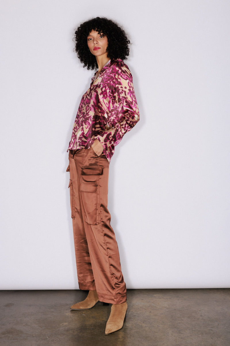 A woman standing against a white plain background, wearing a Catherine Gee Daria French Cuff Silk Blouse Fuchsia Feline features a vibrant pink and purple pattern paired with brown cargo pants with multiple pockets and her hands are in the pockets of the pants and tan suede ankle boots. It highlights the fashion and design elements of the clothing.