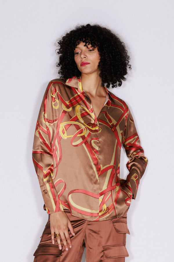 A woman with her one hand in a pocket wears a Catherine Gee Daria French Cuff Silk-Lexington Equestrian blouse features a shiny, silky fabric with a brown base color and an intricate pattern of red and gold equestrian-themed designs paired with brown cargo-style pants with pockets. The background is plain white, which helps to highlight the details of the blouse.