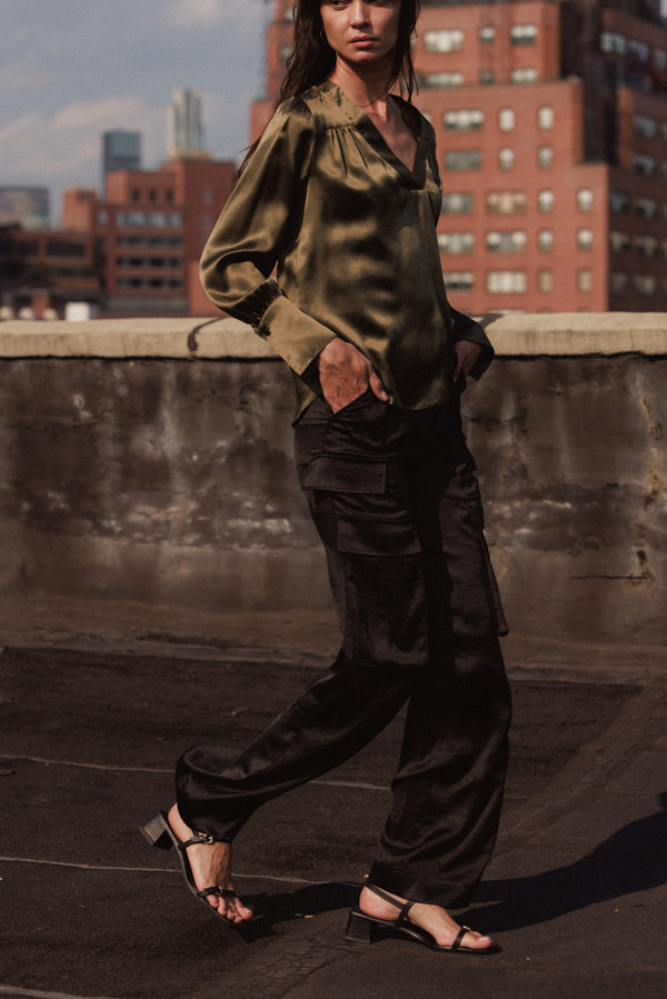 A woman is walking on a rooftop wearing Catherine Gee's black cargo pants paired with a silky olive green blouse and black heeled sandals. The background shows a cityscape with various buildings, adding an urban feel.