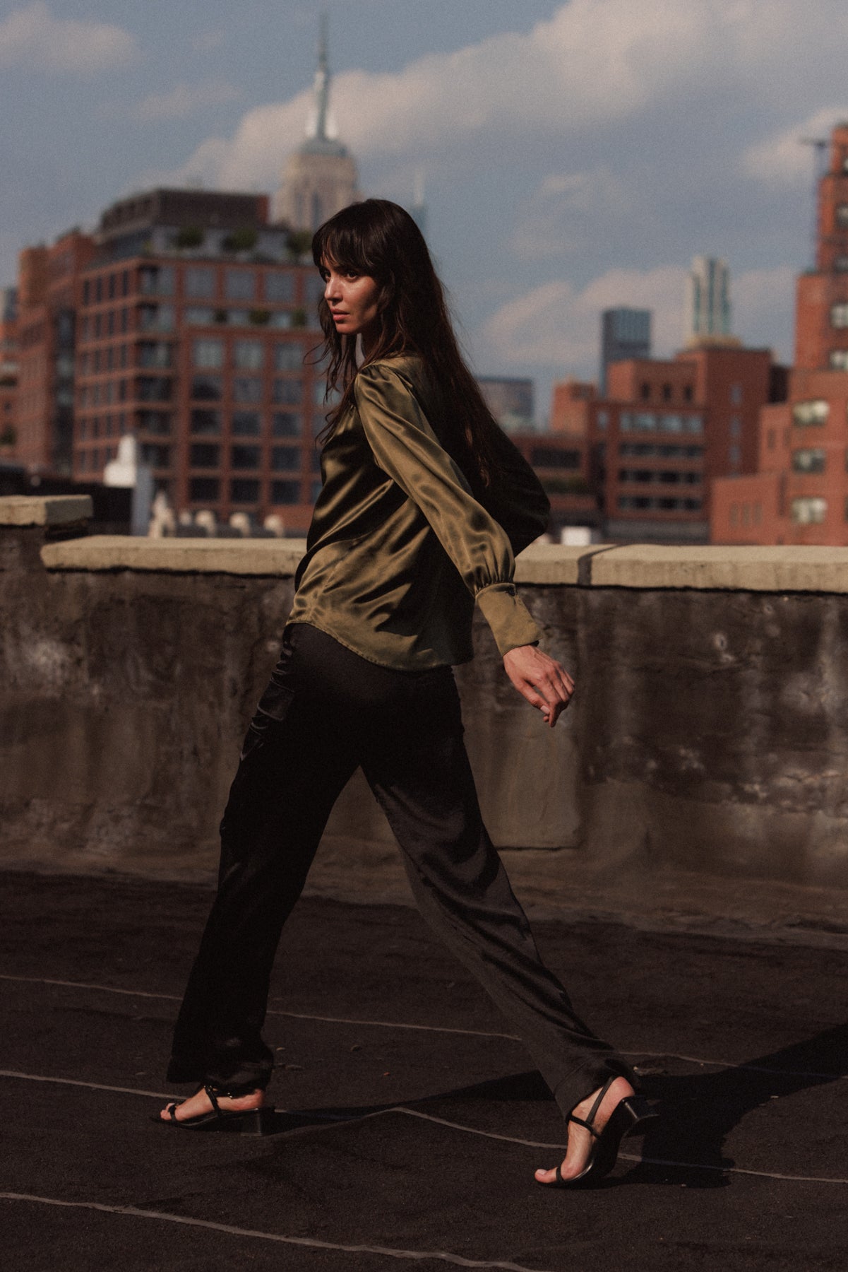 A woman is walking on a rooftop wearing black cargo pants by Catherine Gee paired with a green satin blouse, which captures the fluidity and movement of the outfit. The background features a cityscape with various buildings, adding an urban feel to the scene.