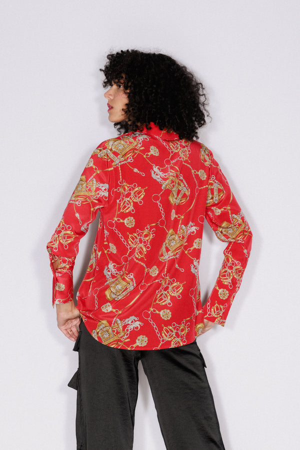 A woman shown from the back wearing a Catherine Gee Daria French Cuff Silk Blouse Faena Red. The blouse is predominantly red with an intricate pattern featuring gold chains and other decorative elements. She has curly hair and also wears dark-colored pants on a plain white background highlights the design and fit of the blouse from the back.