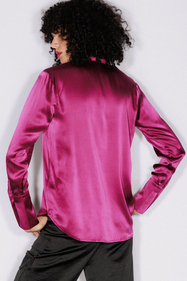 A woman wearing a Catherine Gee Daria French Cuff Silk-Fuchsia blouse features a shiny, smooth texture and a high collar. She is standing with her hands on hips, facing away from the camera, showcasing the back of the blouse on a plain white background, which highlights the vibrant color and texture of the blouse.