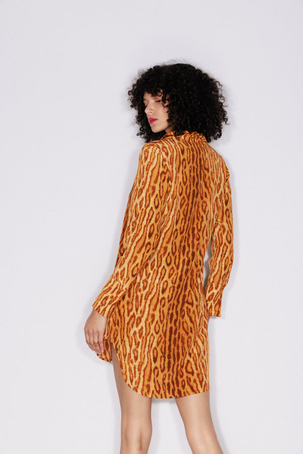 A woman wearing a Catherine Gee Warm Leopard Daria Dress features long sleeves and a loose, flowing fit, and she is standing with her back to the camera, showcasing the dress design. The background is plain and light-colored, highlighting the dress.