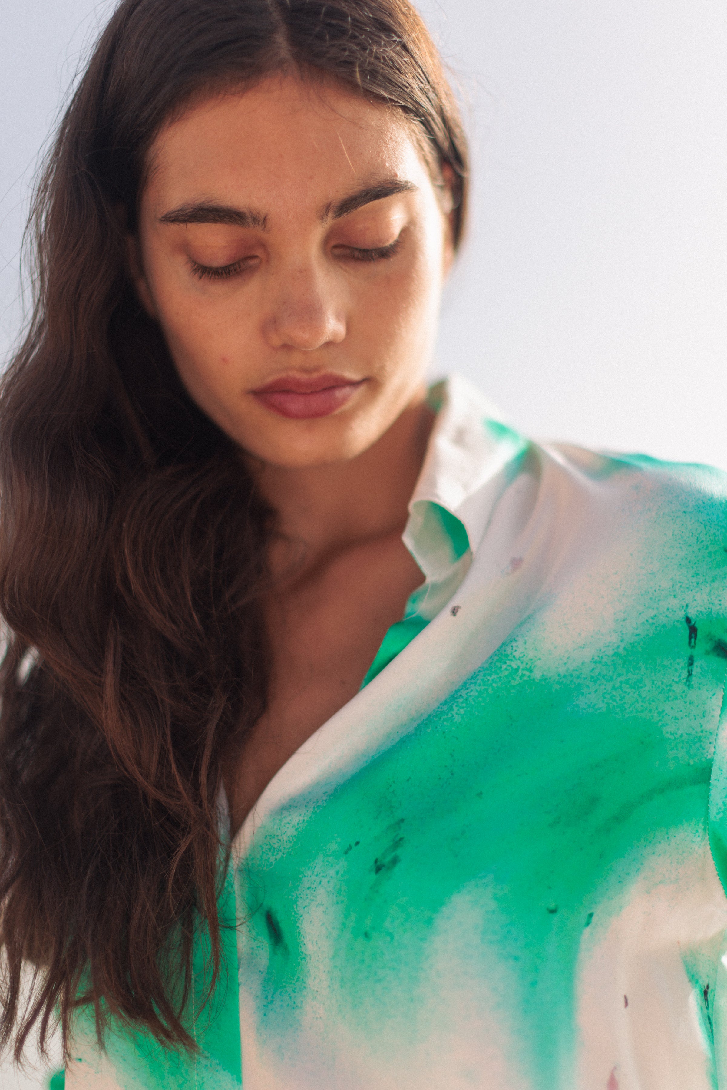 A woman wearing a Catherine Gee Daria French Cuff Silk Teal Art blouse features a vibrant teal color with an artistic, abstract pattern focusing on the upper part of the blouse, highlighting its unique design and texture. The fabric appears to be smooth and silky, reflecting light softly. The collar is slightly open, and the blouse has a relaxed fit, giving it an elegant yet casual look.