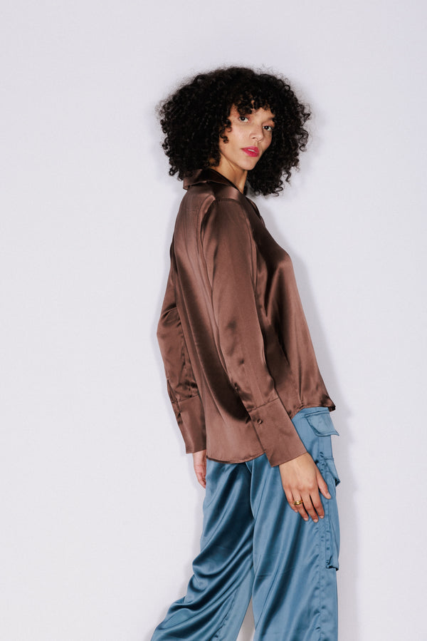 Woman wearing a Catherine Gee Daria French Cuff Silk Blouse in a coffee color. The blouse has a smooth, shiny texture and long sleeves with French cuffs. The person is also wearing loose-fitting blue pants with pockets on the sides.