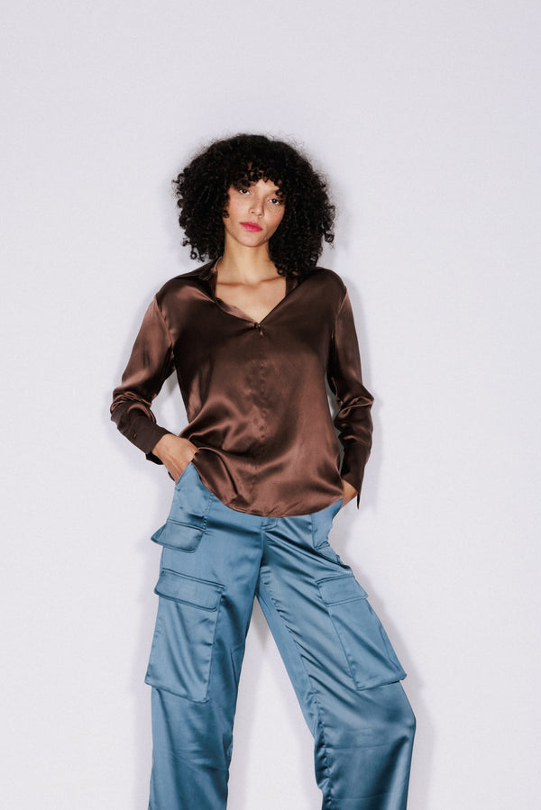 A woman is wearing a Catherine Gee Daria French Cuff silk blouse in a coffee color that features a shiny, smooth texture, a relaxed fit, long sleeves, and a V-neckline. She pairs it with light blue cargo pants that have multiple pockets. The outfit stands out against a plain white background.