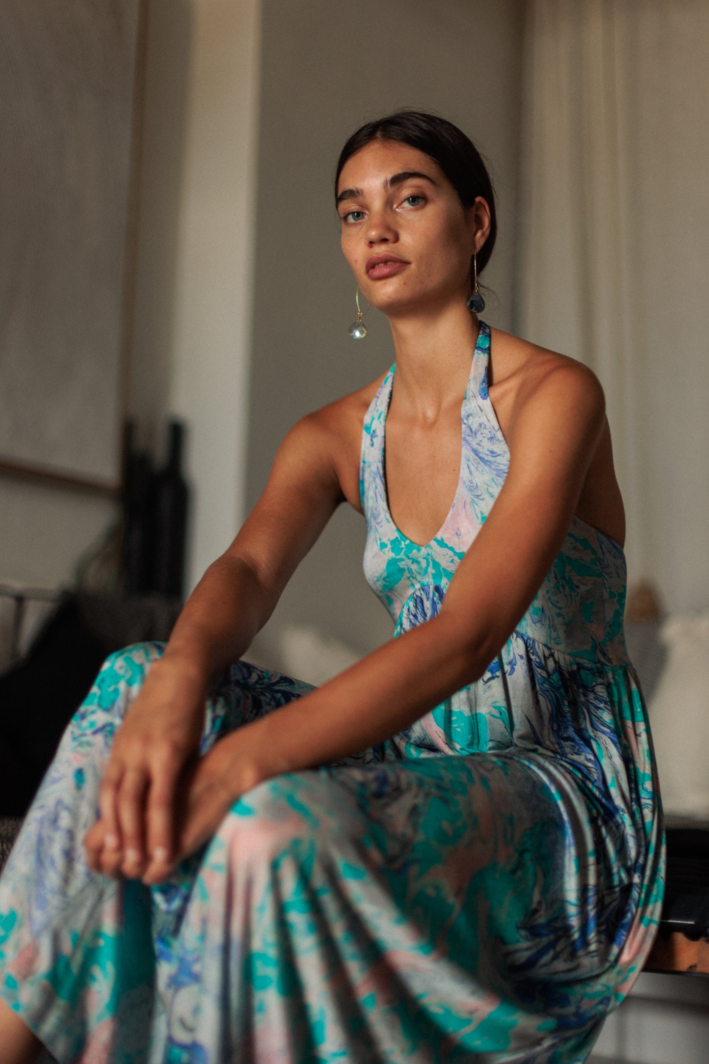 A woman is wearing a Catherine Gee Anna Halter Dress - Cool Swirl features a halter neckline and a vibrant, swirling design in shades of blue, green, and pink. The woman is sitting indoors on a couch, with a background that includes subtle decor elements, ensuring the dress remains the focal point of the image.