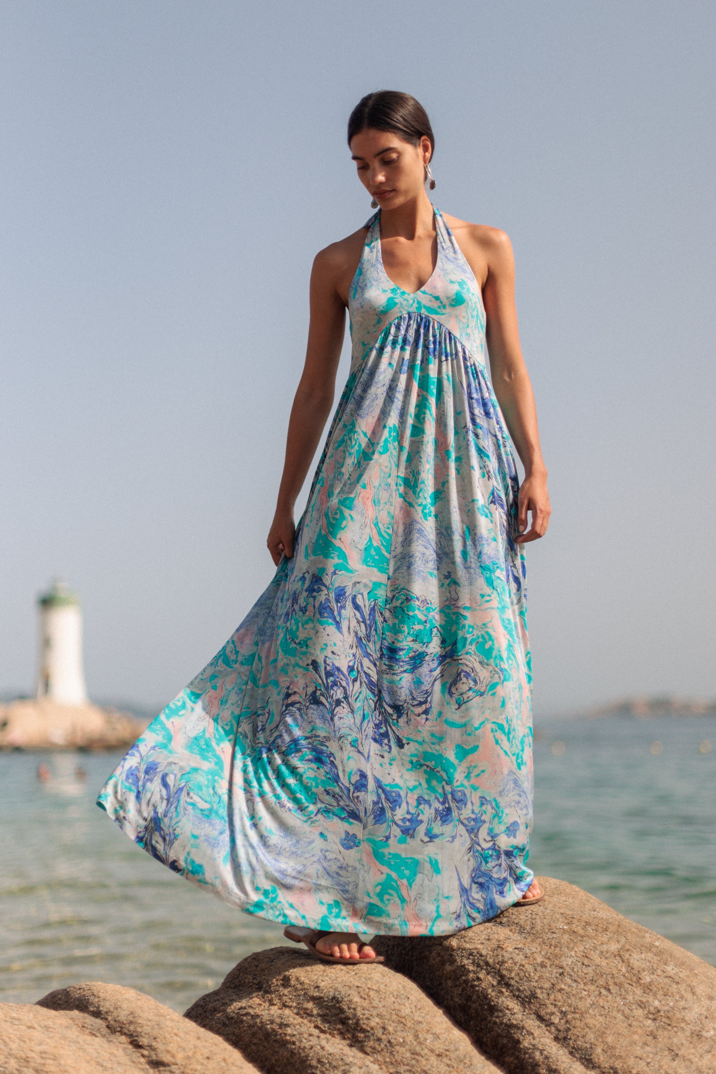 A woman is standing on rocks near the sea, wearing a Catherine Gee Anna Halter - Cool Swirl Dress. The dress features a flowing, floor-length design with a halter neckline and a vibrant blue, green, and pink swirl pattern. In the background, there is a lighthouse and the ocean, adding a scenic and serene element to the image.