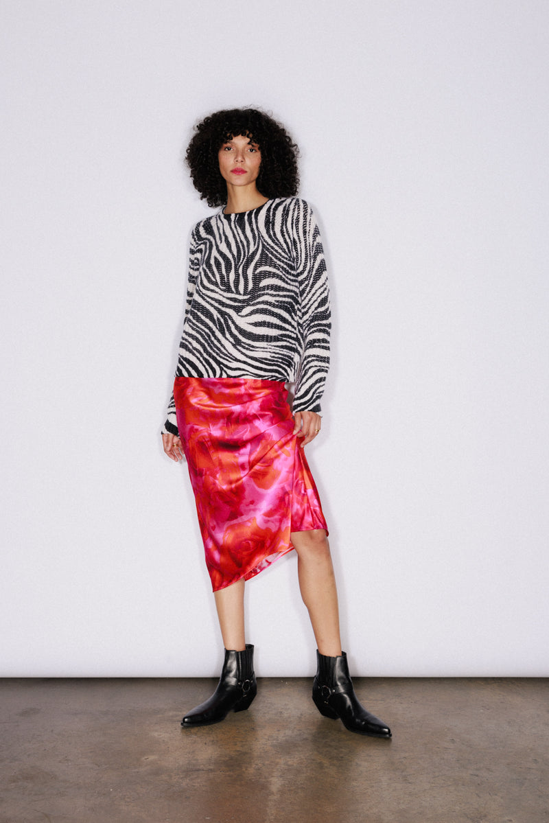 A woman is wearing the Catherine Gee Isabela Chunky Cashmere Zebra sweater, which features a bold black and white zebra print pattern. Made from luxurious chunky cashmere, it provides warmth and comfort. She pairs it with a vibrant red and pink skirt and black ankle boots creating a visually striking and fashionable look that truly stands out.