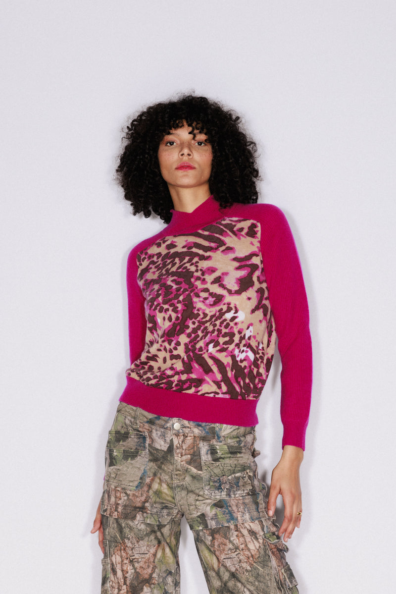 A woman wearing a Catherine Gee Gwen Color Block Cashmere Fuchsia Feline Sweater. This luxurious sweater features a vibrant fuchsia hue with a striking feline print on the front. The bold design is complemented by solid fuchsia sleeves and back, creating a chic contrast. The combination of cashmere fabric and eye-catching design makes this sweater a stylish and comfortable addition to any wardrobe.