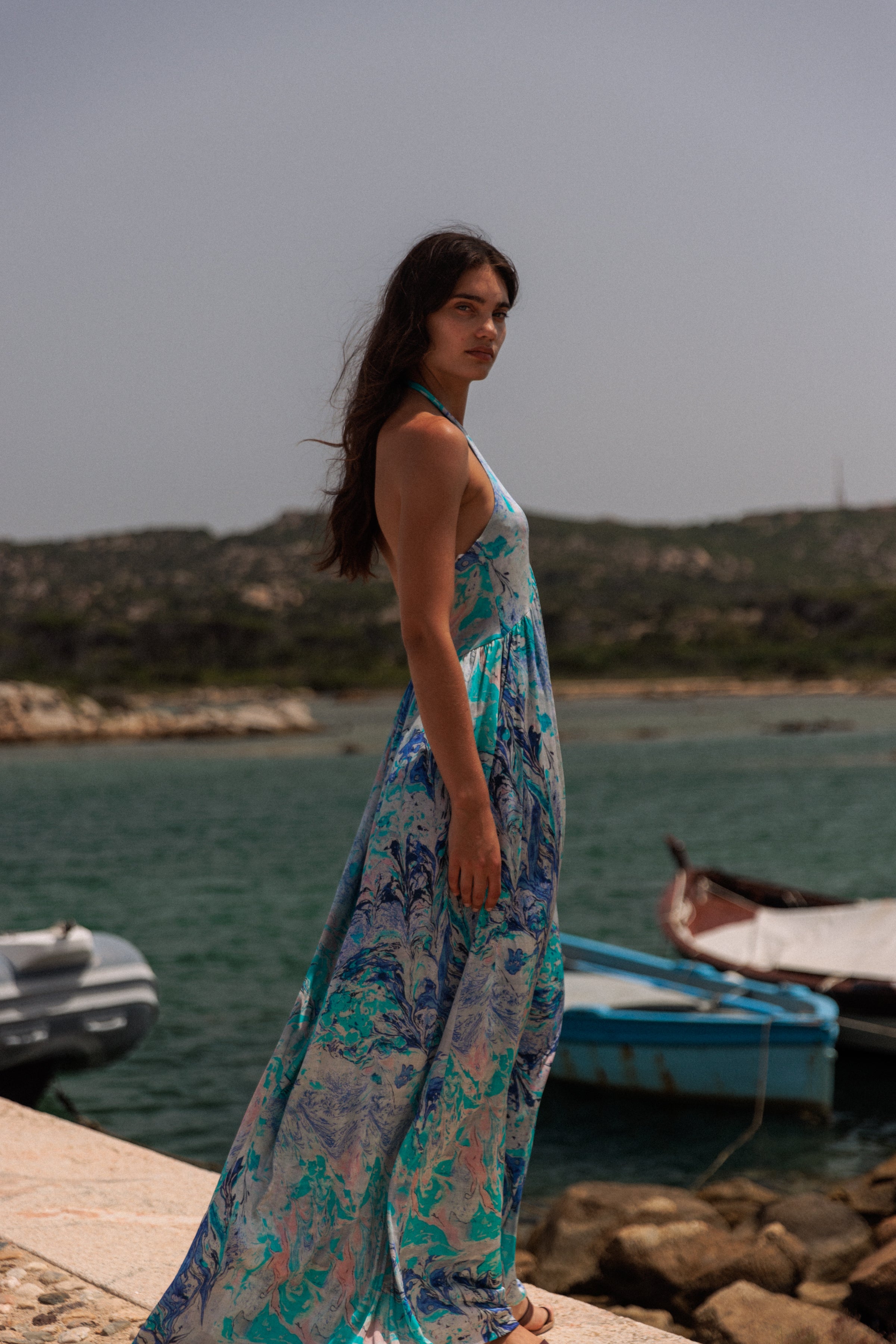 A woman is standing on a stone pathway in the background are boats docked in the water and a hilly landscape, wearing a flowing, sleeveless dress with a halter neckline of the Catherine Gee Anna Halter Dress - Cool Swirl. The dress features a cool swirl pattern in shades of blue, green, and pink. The scene appears to be set in a coastal area, giving a serene and picturesque vibe.