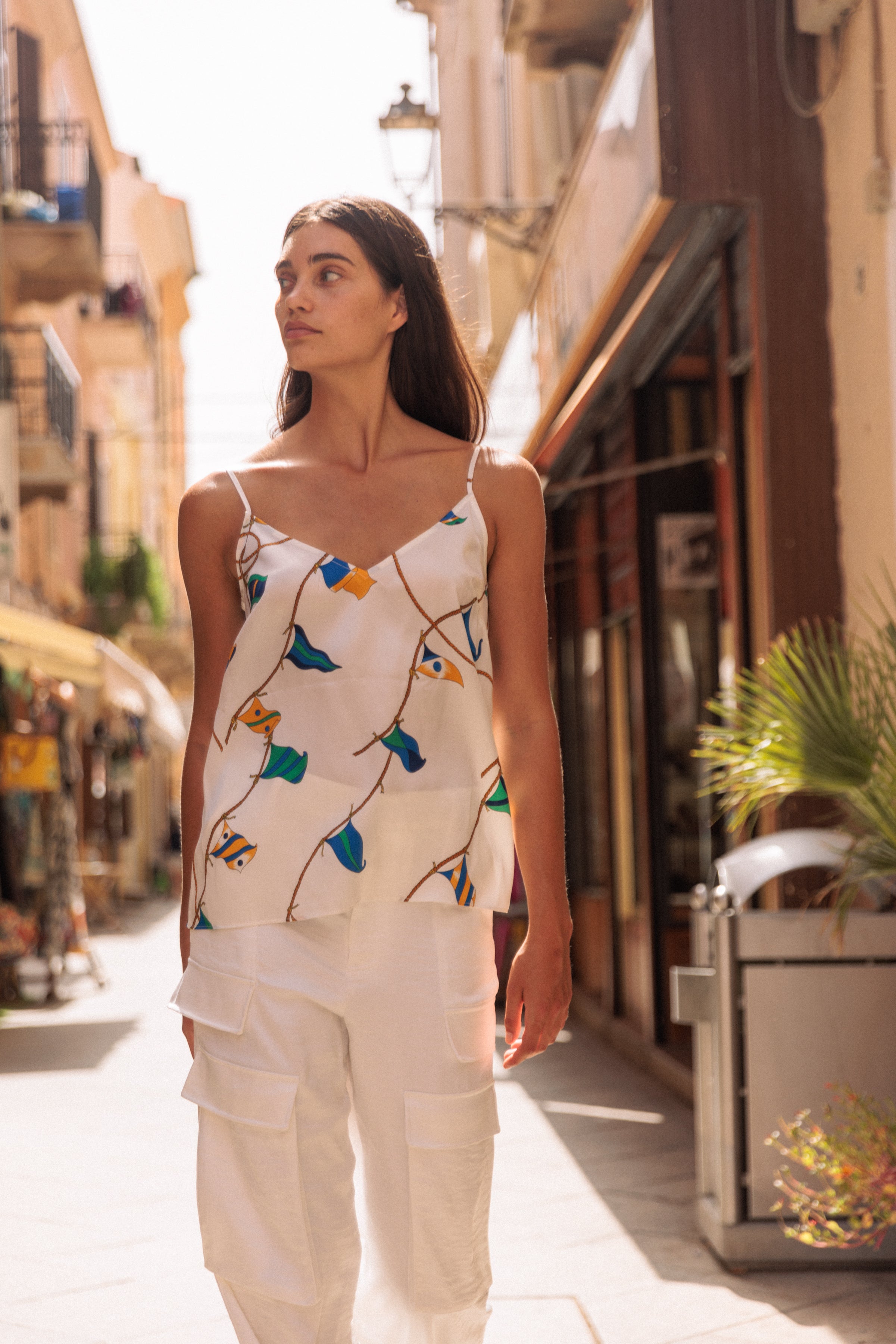 A woman wearing the Catherine Gee Franka Lisbon Sails Cami. This sleeveless top has thin straps and features a colorful sailboat pattern, paired with white pants with multiple pockets. She is walking down a sunlit street lined with shops and buildings, creating a casual, stylish summer look. The vibrant pattern and light fabric make the cami perfect for warm weather.